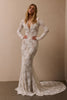 A woman modeling a long-sleeve lace mermaid-style wedding gown with a deep V-neckline and a long train, standing against a plain beige studio wall_XS_