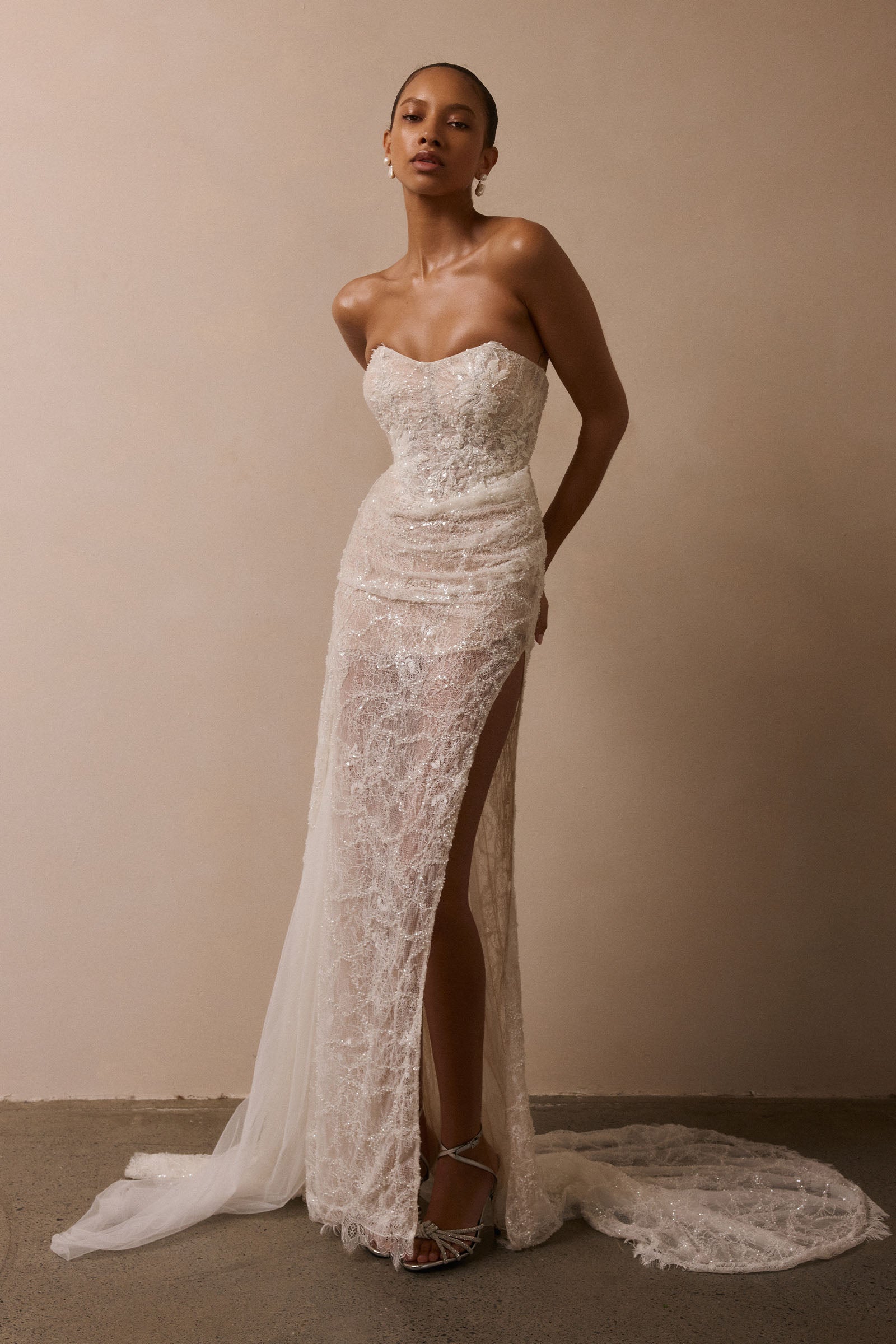 A woman models a strapless lace wedding gown with a sweetheart neckline, fitted bodice, high thigh slit, and a long sheer train against a plain beige studio backdrop_XS_