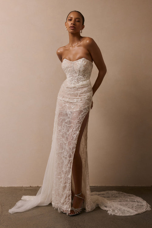 A woman models a strapless lace wedding gown with a sweetheart neckline, fitted bodice, high thigh slit, and a long sheer train against a plain beige studio backdrop_XS_