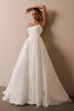 A woman modeling a strapless white A-line wedding gown with a textured lace bodice and a full flowing skirt, posing against a plain beige wall_XS