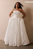 A woman modeling a strapless white ball gown with a structured bodice and voluminous skirt, standing against a warm beige wall_L_