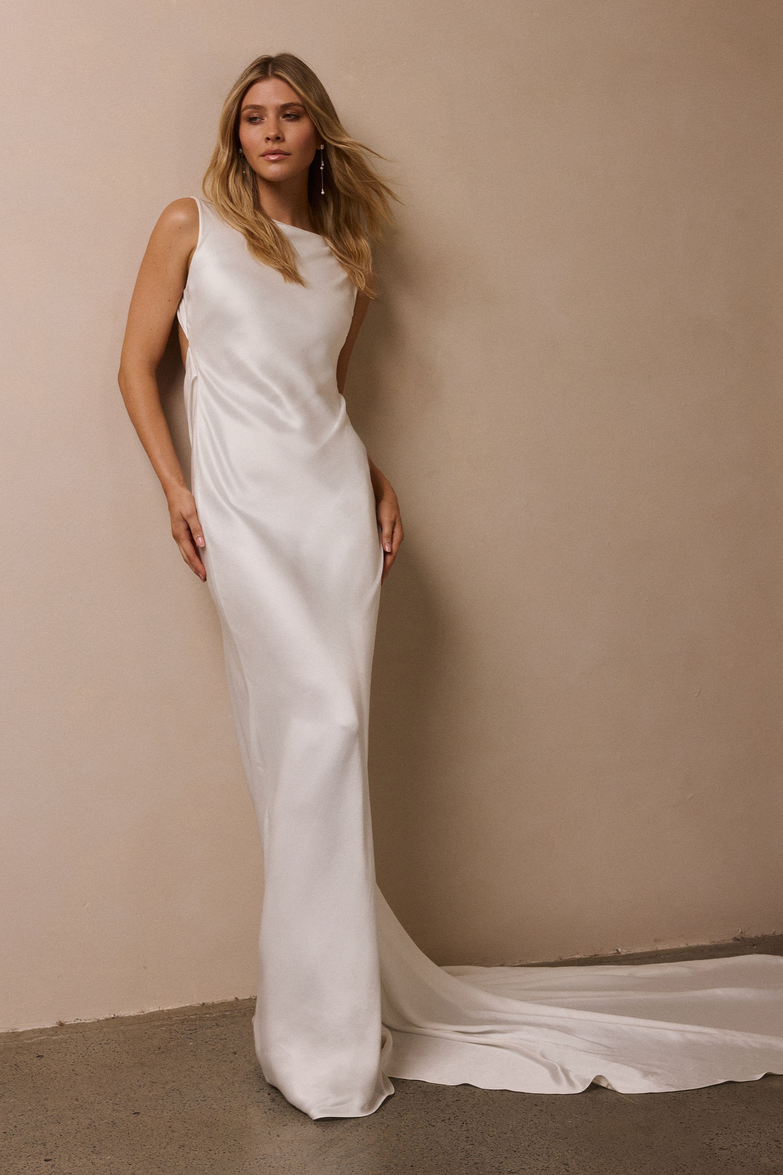 A woman modeling a sleeveless white satin floor-length gown with a high neckline and a long train, standing against a plain beige wall_XS_