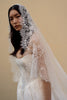A woman wearing a white lace bridal veil and a white wedding dress, photographed against a plain warm beige studio background.