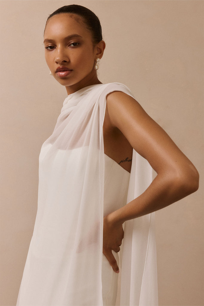 A woman wearing a flowing white draped dress or wrap against a plain beige studio background.