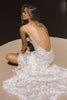 A woman seated on a circular beige platform, photographed from behind, wearing a white lace bridal gown with thin straps, a deep open back, and delicate chain detailing across the upper back.