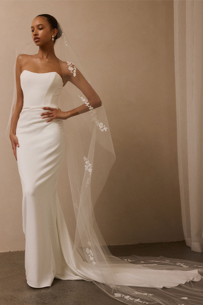 A woman modeling a strapless white wedding gown with a long sheer veil featuring floral lace applique, posed against a warm beige wall.