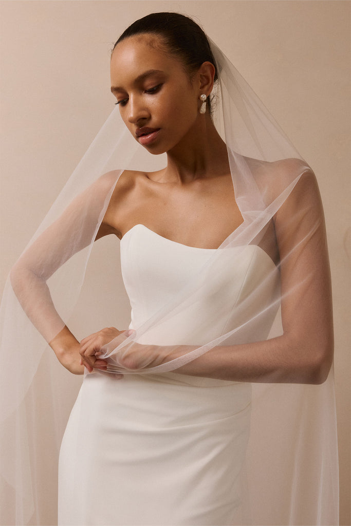 A woman wearing a strapless white wedding dress and a sheer white veil, posed against a warm beige studio background.