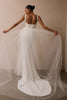 A woman wearing a white wedding dress is photographed from behind, holding out the sheer tulle train against a plain beige studio wall_XS_