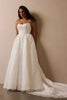 A woman wearing a strapless white wedding gown with a fitted bodice and full lace-trimmed skirt, standing against a plain beige wall_L_