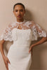 A woman modeling a white sleeveless dress with a sheer white lace bolero-style shrug over the shoulders, posed against a plain beige studio wall.