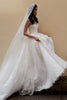A woman wearing a strapless white lace wedding gown with a long sheer veil, posed against a plain beige wall.
