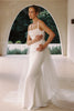 A woman modeling a fitted white wedding dress with thin straps, a draped neckline, and a long sheer tulle overskirt, posed in front of an arched architectural opening.