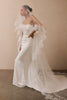 A woman modeling a strapless ivory wedding gown with layered ruffle details, a long sheer veil with lace floral applique posed against a warm beige studio wall.