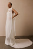 A woman models a floor-length white draped gown with a high neckline and a long flowing train against a plain beige wall and dark floor.