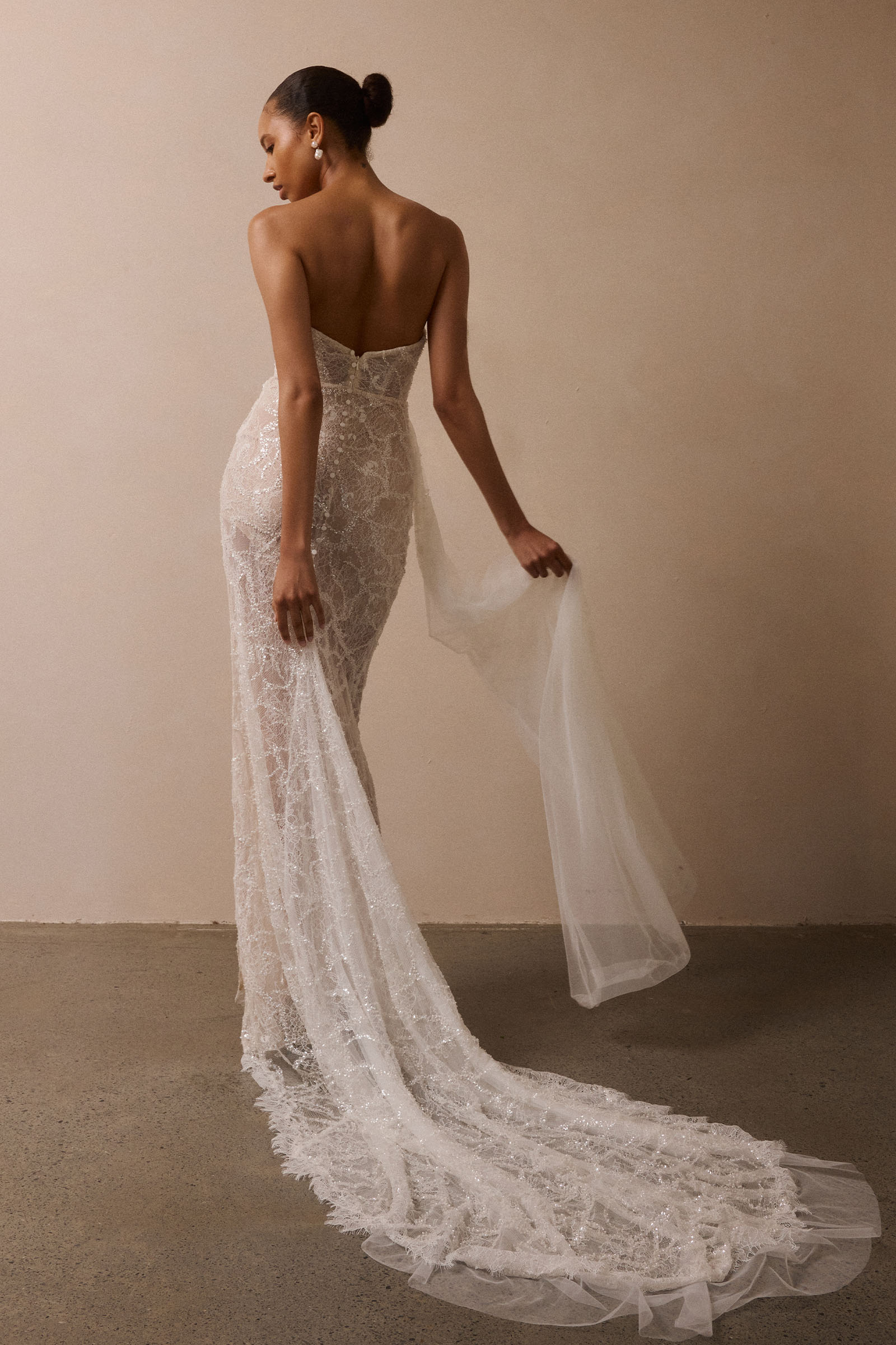 A woman wearing a strapless lace wedding gown with a long sheer tulle train, photographed indoors against a plain beige wall and concrete floor, seen from the back_XS_