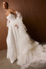 A woman modeling a dramatic white wedding gown with a strapless bodice, sheer long sleeves, and a layered ruffled overskirt and train, posed against a dark wood-paneled wall.