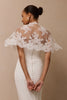 A woman shown from the back wearing a white wedding dress with a sheer lace cape-style back overlay and a row of small back buttons, against a plain beige studio background.