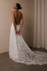 A woman wearing a white lace bridal gown with a low open back and long train, photographed indoors against a beige wall and curtain.