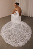 A woman wearing a white lace bridal gown sits on a concrete floor, holding up the back of the dress to show the open back. The dress features intricate floral lace and a long, dramatic train.
