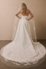 A woman viewed from behind wearing a strapless white wedding gown with a long sheer veil featuring floral lace applique, standing indoors against a plain beige wall.