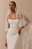 A woman wearing a strapless ivory wedding dress with a sheer lace bridal veil draped over her shoulders, posed against a solid beige studio background.