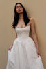 A woman wearing a strapless white lace wedding gown with a fitted bodice and full skirt, standing against a plain beige wall.