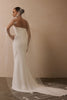 A woman modeling a strapless white wedding gown from the back, wearing a long sheer veil with floral lace applique and a matching headpiece, against a warm beige wall.