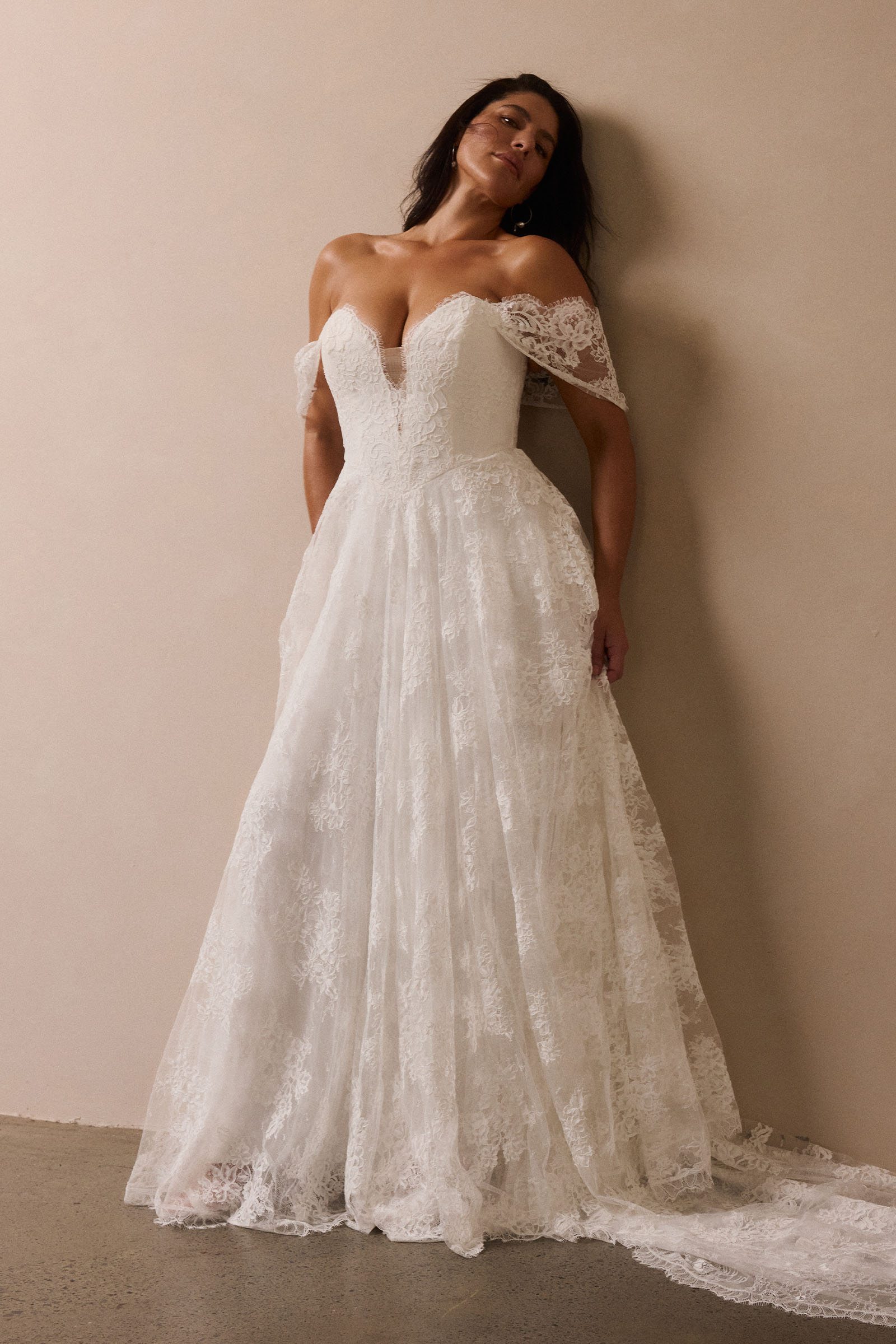 A woman wearing a white off-the-shoulder lace wedding gown with a deep V neckline and a full layered skirt_L_